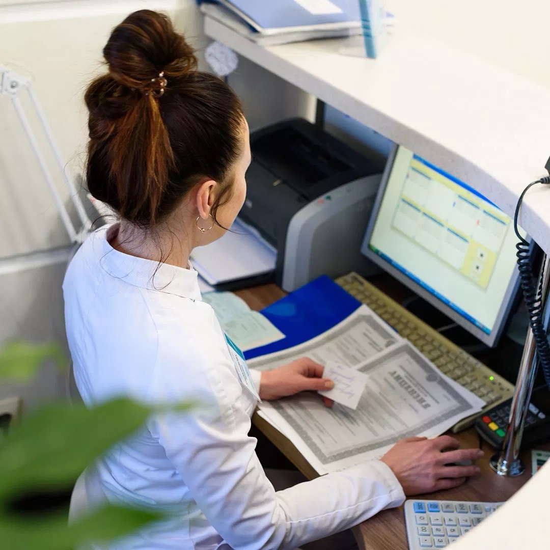 The image shows a woman sitting at a desk with a computer monitor, keyboard, and mouse, working on a document. She appears focused on her task.