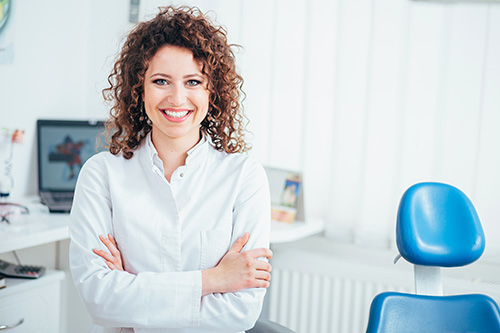 A woman with curly hair, wearing a white shirt and black pants, stands confidently in an office setting with a blue chair and desk in the background.