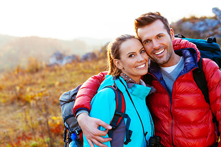 The image shows a man and woman embracing outdoors, with both wearing backpacks, standing on a field during sunset.