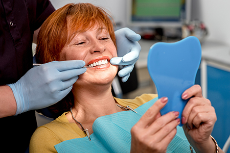 A woman sitting in a dental chair, smiling at a blue toothbrush-shaped object held up by a person wearing a white coat and gloves, with another person adjusting her teeth.