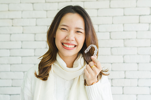 A woman holding a white object with her mouth, smiling, against a brick wall background.