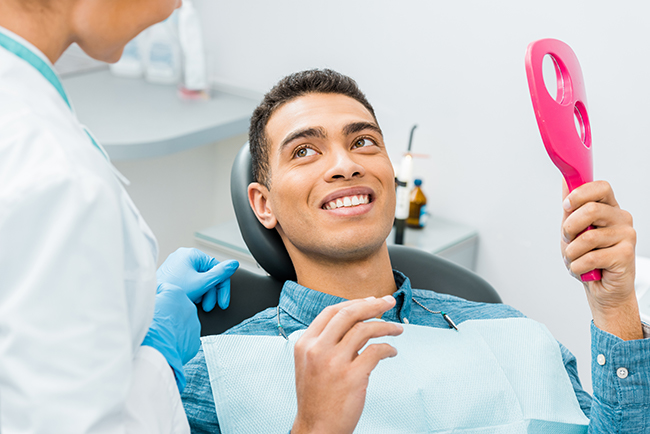 The image shows a smiling man sitting in a dental chair with a pink oral hygiene tool in his hand, being attended to by a dental professional who appears to be performing a cleaning procedure.
