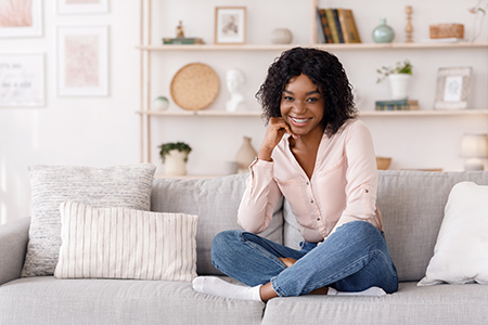 A woman with short hair sits on a couch, smiling at the camera, wearing a pink blouse and blue jeans.