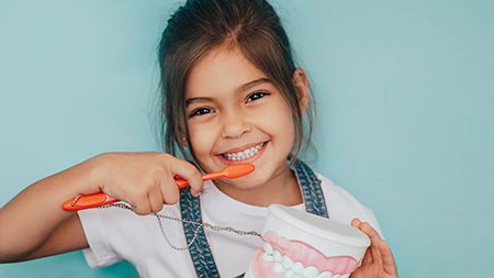 The image shows a young girl brushing her teeth with an orange toothbrush while holding a cup of toothpaste, with one photo on the left showing her smiling and another on the right showing her with her mouth open as she brushes.