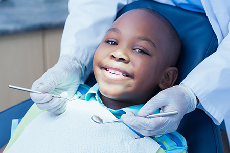 The image shows a young boy sitting in a dental chair, smiling at the camera, with a dentist wearing gloves attending to him, surrounded by dental equipment and tools.
