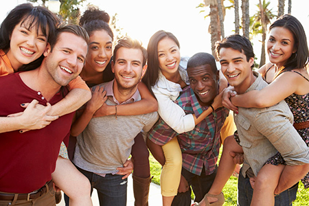 The image shows a group of people posing together outdoors, with a mix of individuals embracing each other, smiling and appearing to be enjoying themselves on what seems to be a sunny day.