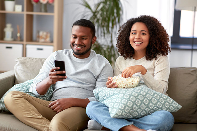 The image shows a man and woman sitting on a couch, smiling at each other while watching something on a phone.